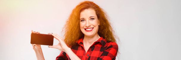 Young red-haired girl in a red checkered shirt. Young girl holding a phone - Photo, Image