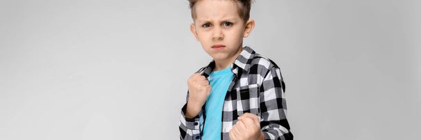 Um menino bonito em uma camisa xadrez, camisa azul e jeans fica em um fundo cinza. O rapaz está na pose de pugilista.
 - Foto, Imagem