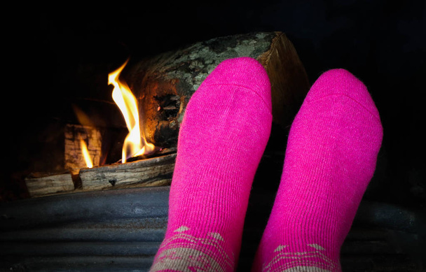 Quemando leña en la chimenea, pies femeninos en calcetines rosados calentándose. Ladrillos de leña en el fuego, calefacción del pie de la mujer
. - Foto, imagen
