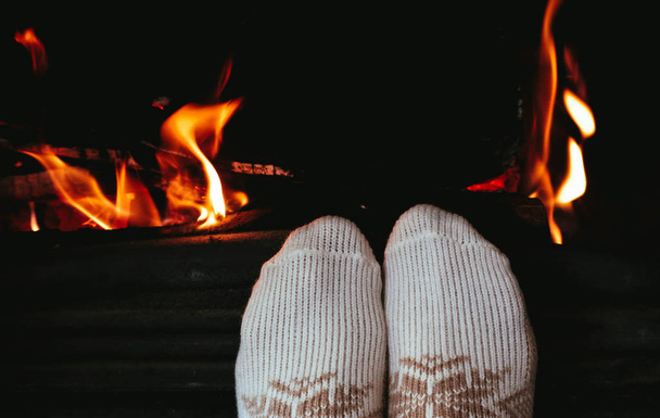 Burning wood at the fireplace, female legs in socks warming up. Firewood bricks at the fire, woman foot heating. - Photo, Image