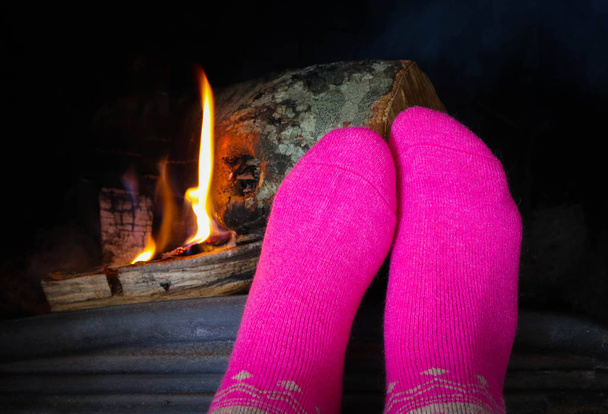 Burning wood at the fireplace, female feet in pink socks warming up. Firewood bricks at the fire, woman foot heating. - Photo, Image