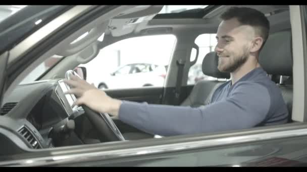 オートサロンで新車で高級車のインテリアを楽しむ男性のクライアント。ホイールを維持、横、カメラ目線、最初の計画に車の警報システムを見せて男を笑っています。購入の概念. - 映像、動画