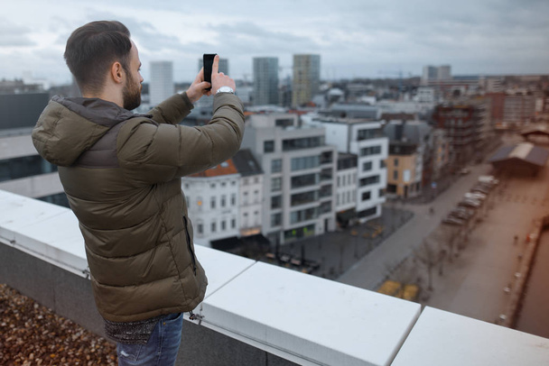 Young hipster taking pictures of the city on a smartphone from the roof of the building. - Fotografie, Obrázek