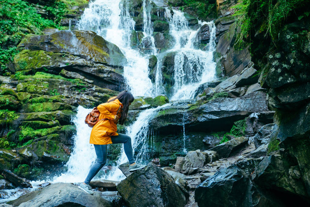 Frau springt im gelben Regenmantel von Felsen zu Felsen im Wald. Wasserfall auf Hintergrund - Foto, Bild