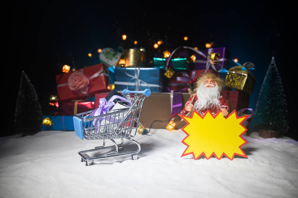 新年やクリスマス商戦のコンセプト。キャンペーンを保存します。サンタ クロースは、雪の上、テキストの黄色いステッカーを保持しています。背景のギフト ボックスのスタック. - 写真・画像