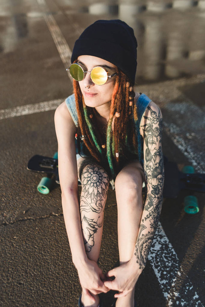 young girl with tattoos and dreadlocks in a blue cap sits on a longboard against the background of concrete structure - Fotoğraf, Görsel