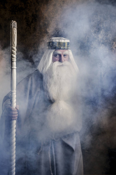 A grey-haired bearded and gloomy wizard in a gray cloak is doing witchcraft in smoke against a magical dark golden textured background. - Foto, imagen