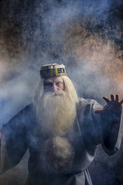 A grey-haired bearded and gloomy wizard in a gray cloak is doing witchcraft in smoke against a magical dark golden textured background. - Foto, imagen