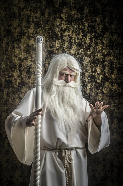 A grey-haired bearded severe wizard in a white cloak and a cap with a staff against a dark golden textured background. - Foto, imagen