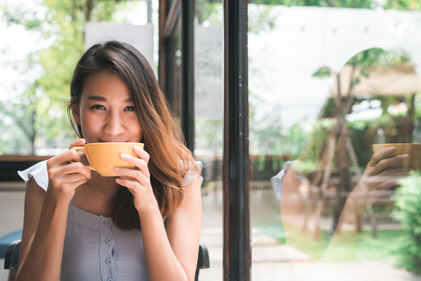 Iloinen aasialainen nuori nainen juo lämmintä kahvia tai teetä nauttien siitä istuessaan kahvilassa. Houkutteleva onnellinen aasialainen nainen, jolla on kuppi kahvia
. - Valokuva, kuva