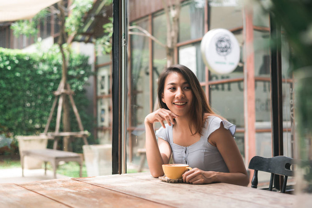 fröhliche asiatische junge Frau, die warmen Kaffee oder Tee genießt, während sie im Café sitzt. attraktive glückliche asiatische Frau mit einer Tasse Kaffee. - Foto, Bild