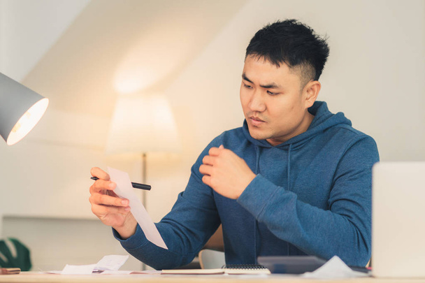 schöne junge lächelnde asiatische Mann arbeitet Laptop auf Schreibtisch im Wohnzimmer zu Hause. asiatischer Geschäftsmann, der im Homeoffice Notizbücher über Finanzen und Rechner schreibt. Zeit zu Hause genießen. - Foto, Bild