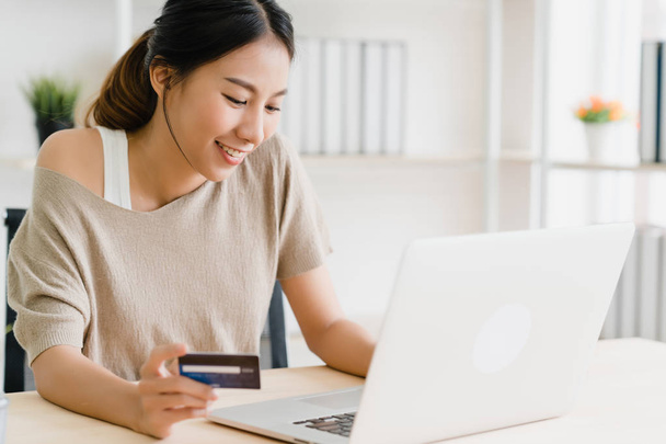 Азиатская женщина, пользуясь компьютером или ноутбуком, совершает онлайн-покупки с помощью кредитной карты, а свитер носит сидя дома на столе в гостиной. Концепция "женщина дома"
. - Фото, изображение