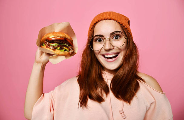 Close up portrait of a hungry young woman eating burger isolated over pink background - Foto, imagen