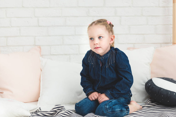 Cute little girl in blue denim overalls sitting on the bed. - Zdjęcie, obraz