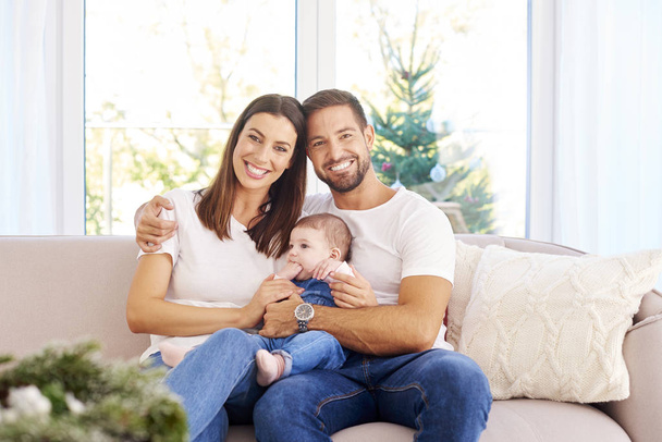 Onnellinen nuori perhe istuu sohvalla vauvansa kanssa ja nauttivat toistensa seurasta jouluna.
.  - Valokuva, kuva