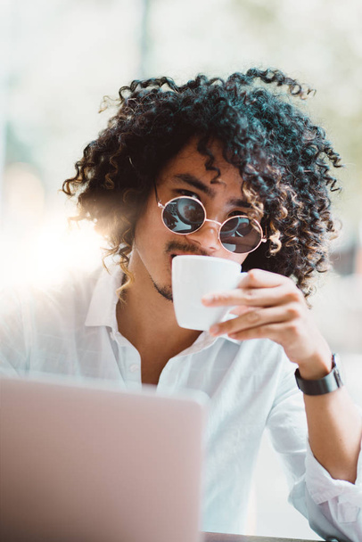 Vertical shot of a young handsome curly male freelancer in sunglasses drinking delicious coffee in an outdoor bar; Asian man entrepreneur drinking tea in a street cafe with a laptop in foreground - Photo, Image