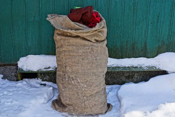 full brown sackcloth bag stands in the snow near the wall of the house - Photo, Image