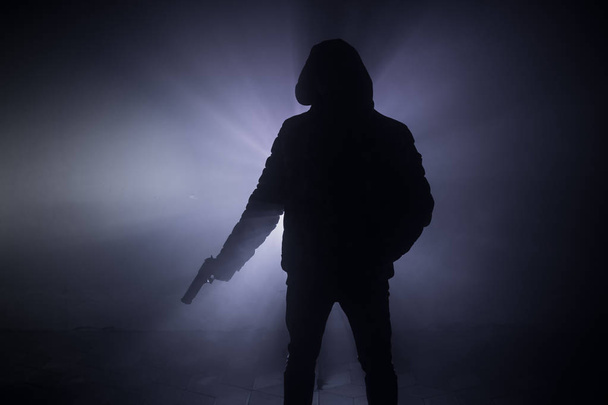 Silhouette of man with pistol ready to attack on dark toned foggy background or dangerous bandit holding gun in hand. Shooting terrorist with weapon theme decor - 写真・画像