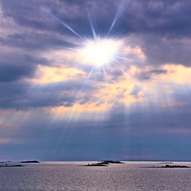 Aurinko pilvien takana valonsäteet loistavat alas mereen
 - Valokuva, kuva