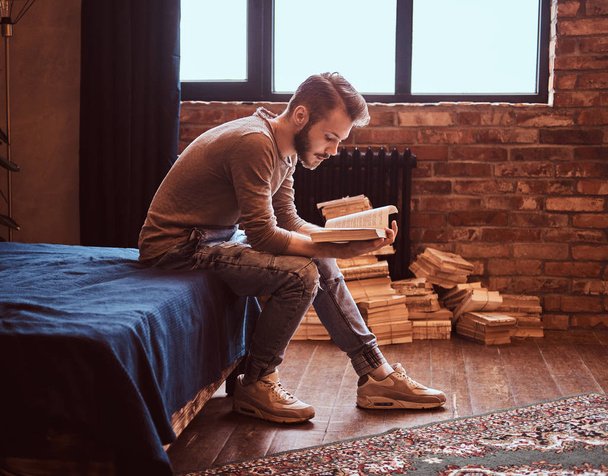 Student met stijlvolle baard en haren is voorbereiden op examens, zittend op het bed en het lezen van een boek. - Foto, afbeelding