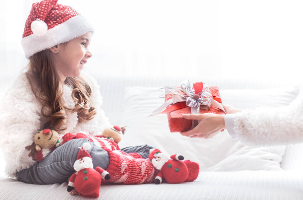 kleines süßes Mädchen mit Weihnachtsgeschenk im Weihnachtsmannkostüm auf Sofa, Weihnachts- und Neujahrskonzept - Foto, Bild