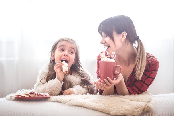Mom and her cute daughter are eating Christmas sweets in the living room on a bright background, the concept of family values and festive atmosphere - Foto, Imagem
