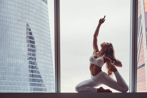 Menina bonita vestindo sportswear fazendo exercício de ioga em uma soleira da janela em um apartamento arranha-céu
. - Foto, Imagem