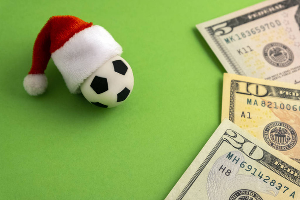 The red Santa Claus hat is dressed on a souvenir soccer ball next to the dollars on a green background. The concept of New Year football. Christmas sports betting to win or lose. - Photo, Image