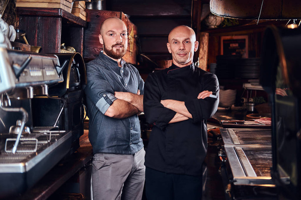 Chef with his assistant posing for a camera with his arms crossed in a restaurant kitchen. - Фото, изображение
