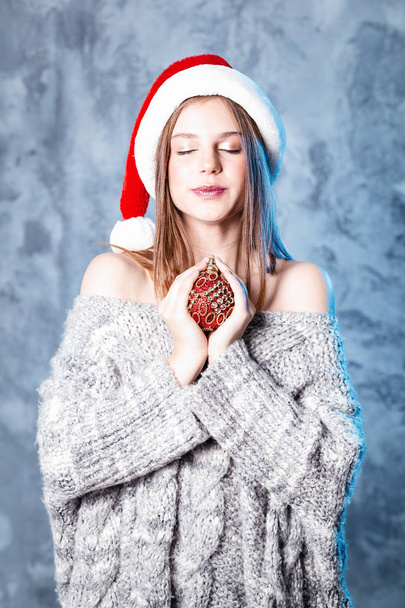 Merry Christmas and happy new year! happy girl with ball toy in hands looks very pleased with closed eyes dreaming. Close portrait on gray background. Girl in santa hat and sweater - Фото, изображение
