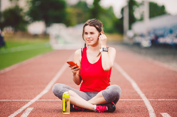 theme healthy lifestyle, sports music. beautiful young Caucasian woman rests sitting treadmill running rubber stadium uses technodogy mobile phone hand insert earphones into ear include music training - Foto, imagen