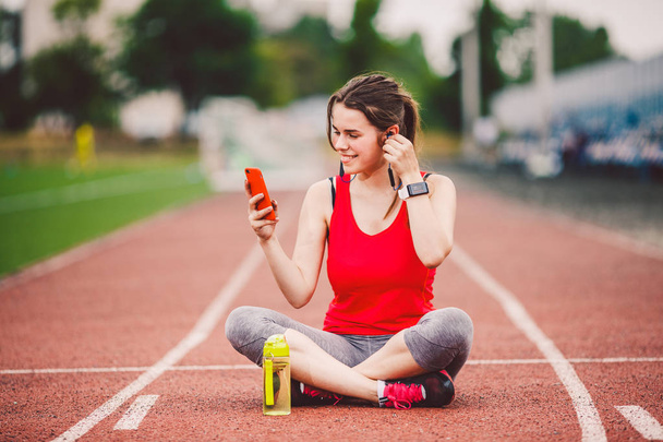 theme healthy lifestyle, sports music. beautiful young Caucasian woman rests sitting treadmill running rubber stadium uses technodogy mobile phone hand insert earphones into ear include music training - Foto, immagini