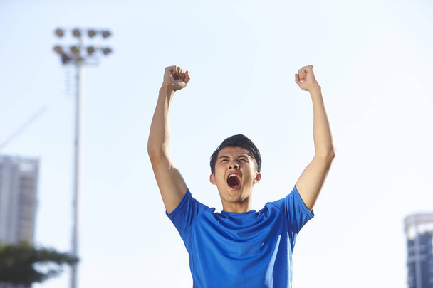 jonge Aziatische mannelijke atleet vieren overwinning met vuisten en armen verhoogd - Foto, afbeelding