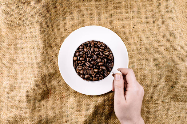 Рука тянется за кружку с кофейными зернами на коричневом, структурированном фоне - Фото, изображение