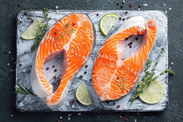 Two raw fresh salmon or trout steaks on ice, rich in omega-3 oil, with lime, thyme and olive oil on a dark background. Healthy and dietary food. Top view. Flat lay. - Foto, Imagen