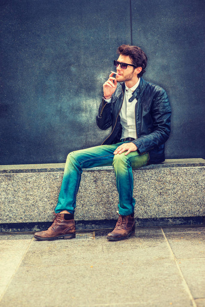Wearing black leather jacket, blue jeans, brown boot shoes, sunglasses, a young guy with beard sitting on marble bench in corner, smoking cigarette during working break, trying mind calming down - Foto, Imagem