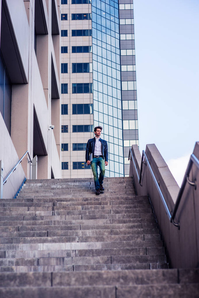 Молодой человек с бородой, в кожаной куртке, джинсах, с ноутбуком на руке, спускающийся по лестнице возле офисного высокого здания после напряженного рабочего дня, идущий домой. Фильтрованный взгляд с фиолетовым оттенком
. - Фото, изображение
