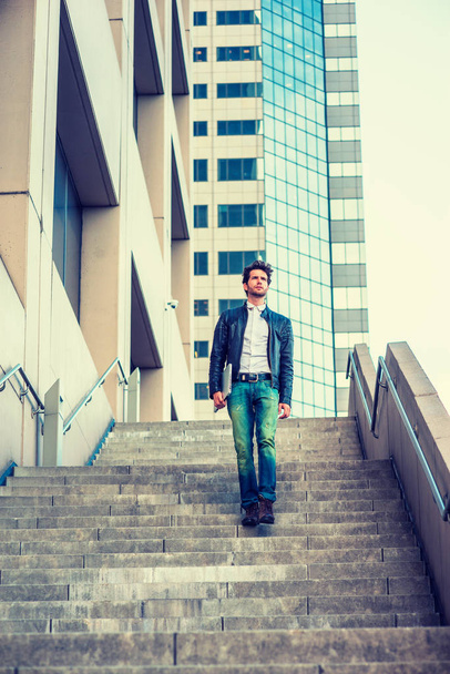 A professional with beard, wearing leather jacket, jeans, arm carrying laptop computer, walking down stairs outside office high building after busy working day, going home. Filtered look. - Foto, Imagem