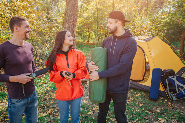 Picture of three friends in forest. Young woman smiles and looks at bearded guy. He holds sleeping bag. Another young man holds knife. - Photo, Image