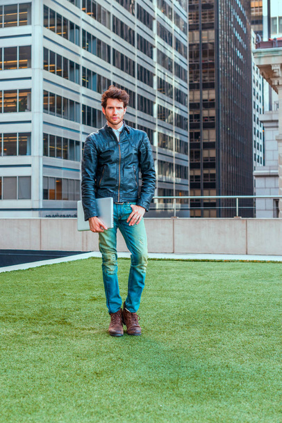 European graduate student studying in New York. Wearing black leather jacket, blue jeans, carrying laptop computer, a young guy with beard, standing on green lawn in business district, thinking. - Photo, Image