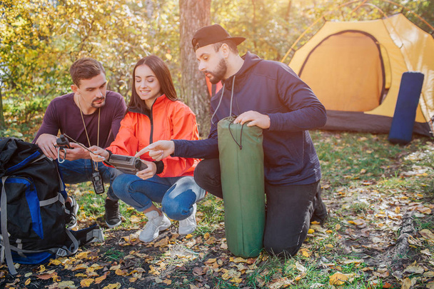 Three friends sit in squad position and look at radio young woman holds. Bearded guy holds sleeping bag. Another young man has knife in cover. They talk. - Photo, image
