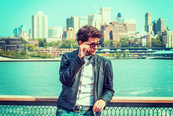 European man with beard traveling in New York, wearing black leather jacket, holding sunglasses, standing by river, narrowing eyes, looking over glasses frame, watching away. Brooklyn on background. - Photo, Image