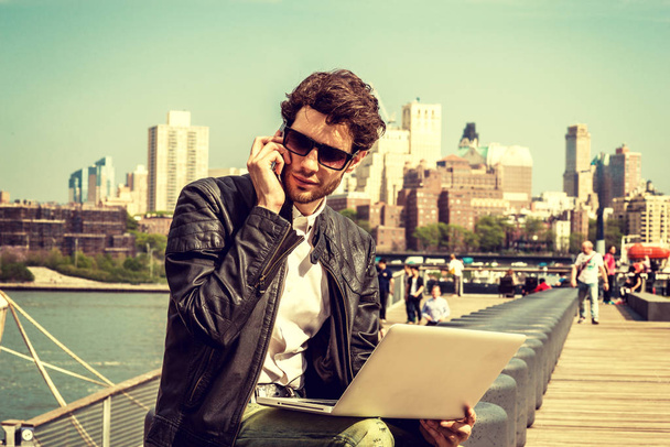 Geschäftsmann auf Reisen, arbeitet in New York. Sonnenbrille auf, ein junger Mann mit Bart, der auf einer Bank am Hafen sitzt, am Laptop arbeitet und gleichzeitig telefoniert. Retro gefilterter Look. - Foto, Bild