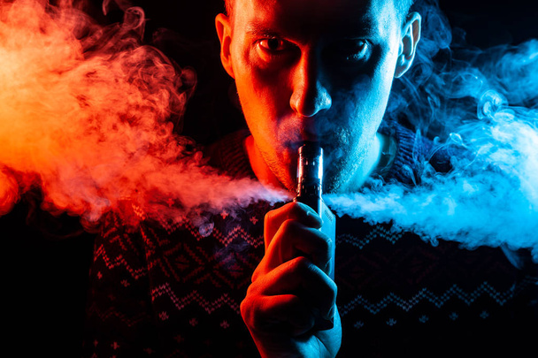 Porträt eines Mannes mit einem Schatten auf einem ernsten Gesicht mit einer farbigen Hintergrundbeleuchtung aus blauem und rotem Rauch, der einen Vape raucht und mehrfarbigen Rauch in verschiedene Richtungen auf einem schwarzen, isolierten Hintergrund ausatmet - Foto, Bild