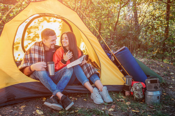 Young peoaple sitting in tent and look at each other. She holds opened book while guy has thermos in hands. She holds thermocup. There backpack with equipment at tent. - Foto, imagen