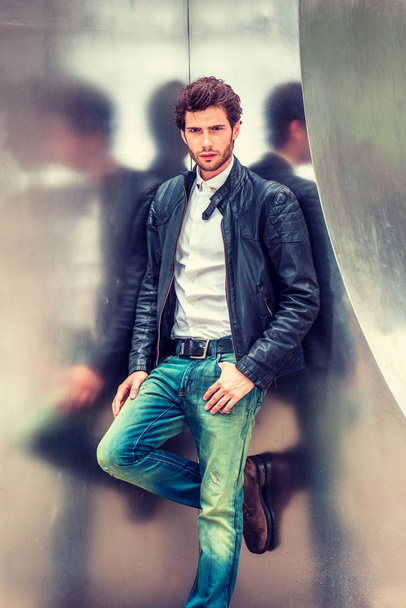Man Spring/Autumn Casual Fashion. Young guy with beard, wearing black leather jacket unbuttoned, blue jeans, leaning against silver metal wall, relaxing, thinking, waiting for you. - Photo, Image