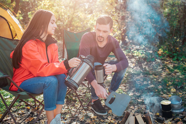 Young woman sit on folding chair and poures hot water from thermos into mans cup. She does it accurate. Guy hold thermocup in hand. They sit at fire together. - Fotoğraf, Görsel