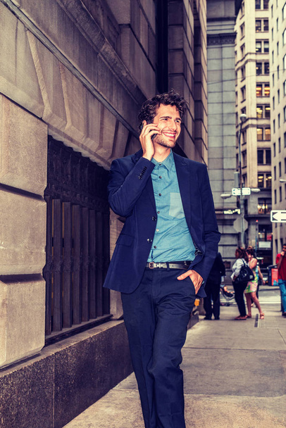 Европейский бизнесмен путешествует по Нью-Йорку. Молодой парень с бородой идет по переполненной высокой улице, разговаривает по мобильному телефону, улыбается, взволнован хорошими новостями. Технологии в повседневной жизни
. - Фото, изображение
