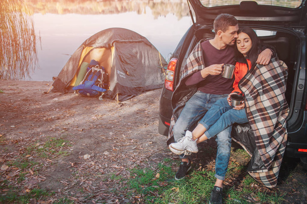 Young man embrace woman. They sit in trunk. Model is covered with blanket. Couple is at lake. There is tent at waterline. - Фото, изображение